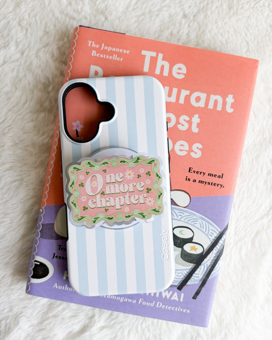 A light blue and white striped phone case with a pink and green 'One more chapter' grip, resting on top of a book on a white faux fur surface.
