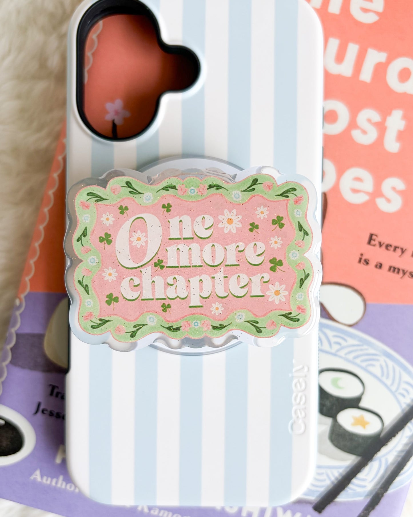 A phone case with a light blue and white vertical stripe pattern, featuring a central pink decorative grip with the text 'One more chapter' in white script, surrounded by small green leaves. The case is displayed on top of books on a white textured surface.
