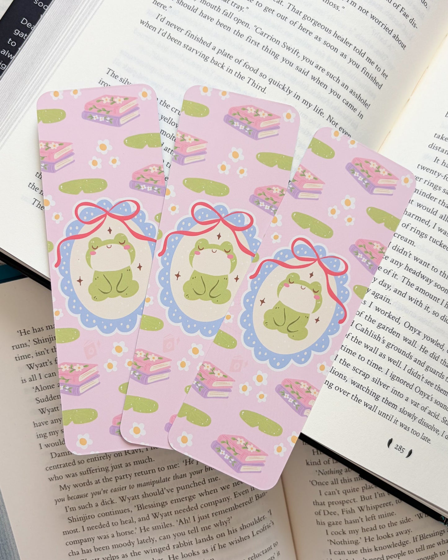 Three pink bookmarks with cartoon frog designs on an open book.