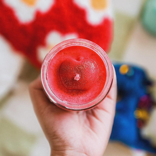 Hand holding red heart-shaped candle in glass jar, glittery wax, bookish merch for readers