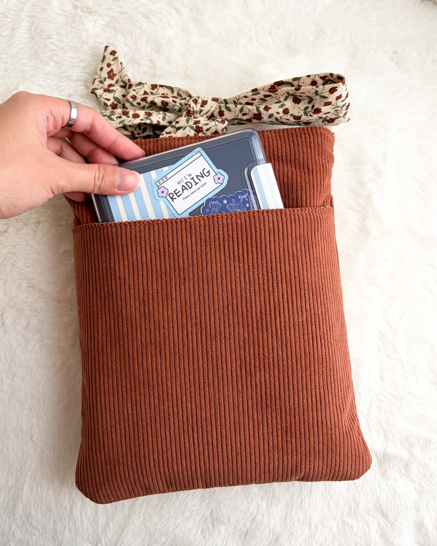 Hand holding e-reader in brown corduroy book sleeve with floral bow on white background