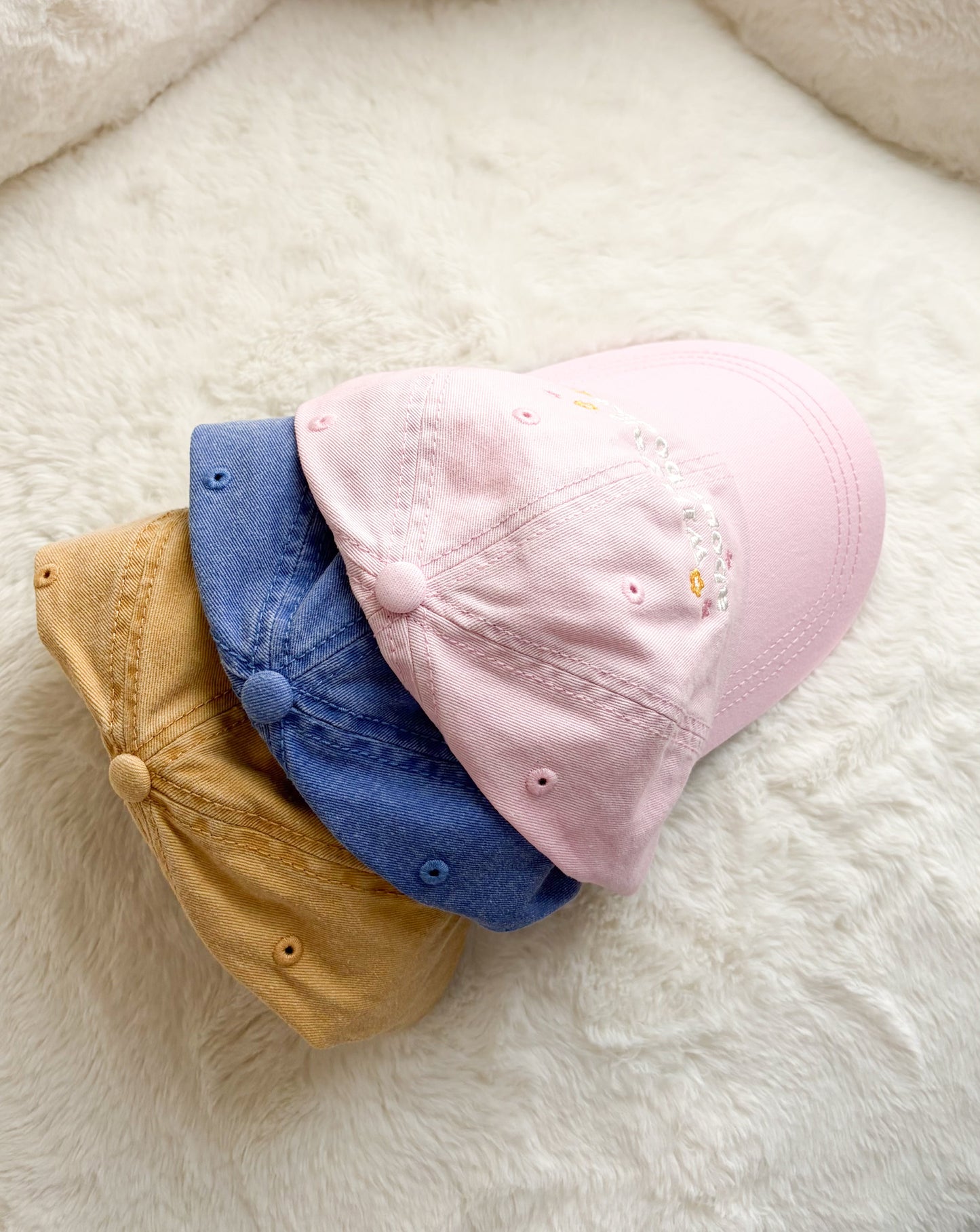 Three pastel baseball caps in pink, blue, and tan on a soft cream background