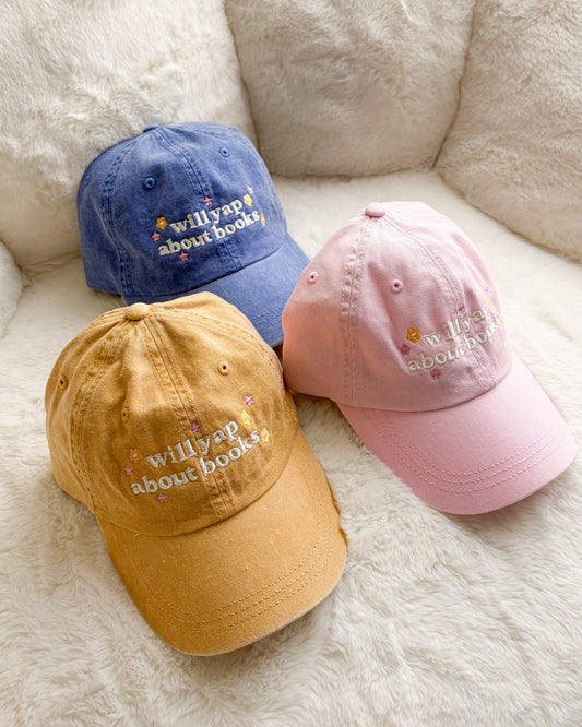 Three embroidered baseball caps in yellow, blue, and pink with 'willyap about books' text.