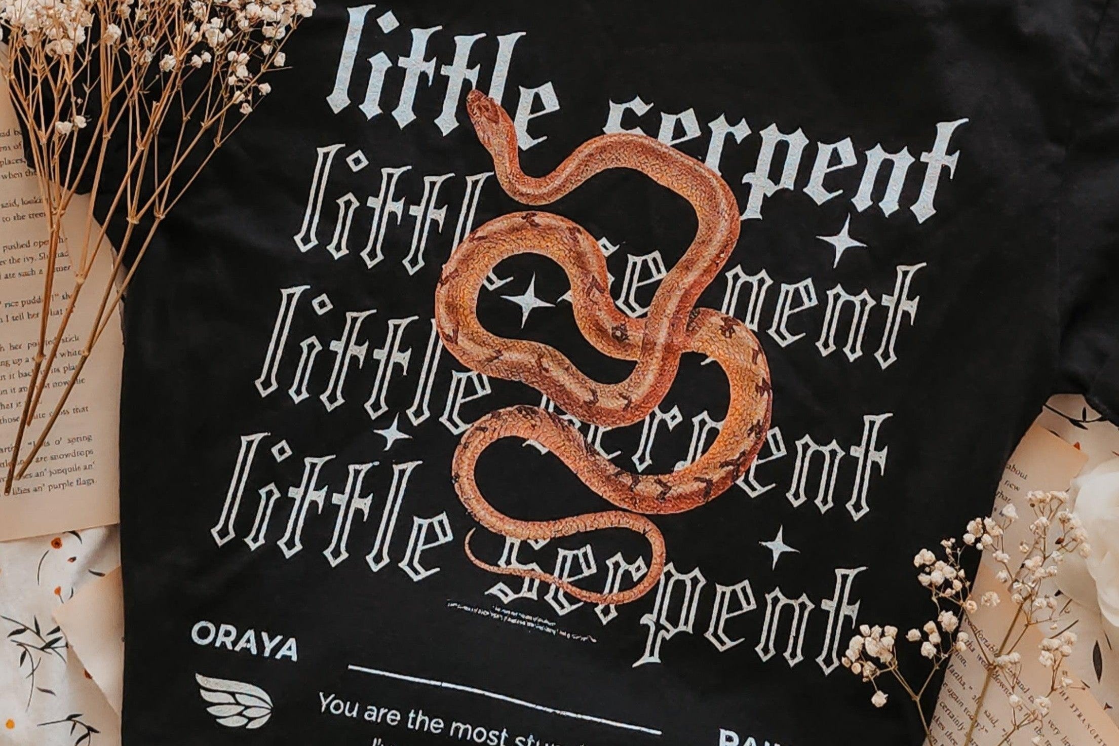 Black shirt with 'little serpent' text, orange snake graphic, dried flowers, and book pages