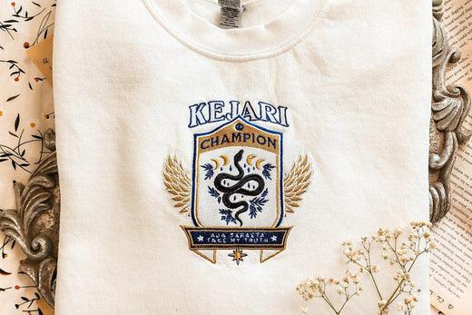 Folded white shirt with 'Keijari Champion' crest, winged snake emblem, book themed apparel