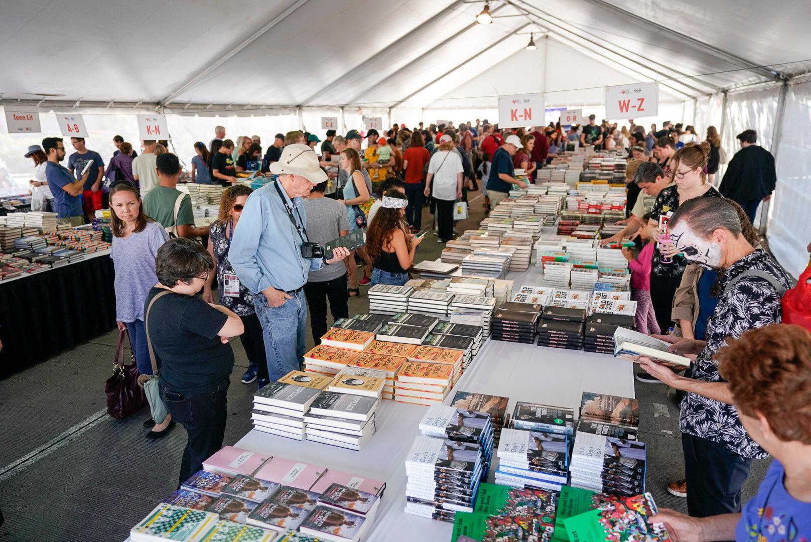 Crowded book event under a tent with people browsing tables full of books