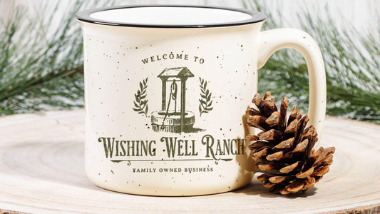 Speckled cream campfire mug with 'Wishing Well Ranch' text, pine cone, and pine branches.