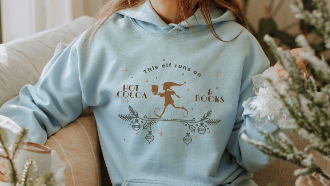 Light blue hoodie with festive elf graphic and 'This elf runs on hot cocoa & books' text