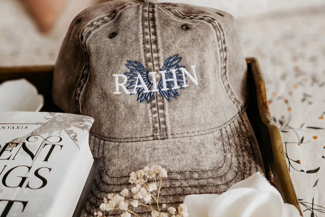Embroidered RAHN gray cap with blue wings design, next to a book and white flowers