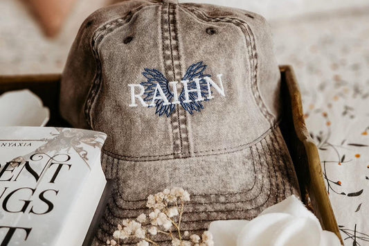 Embroidered RAHN gray cap with blue wings design, next to a book and white flowers