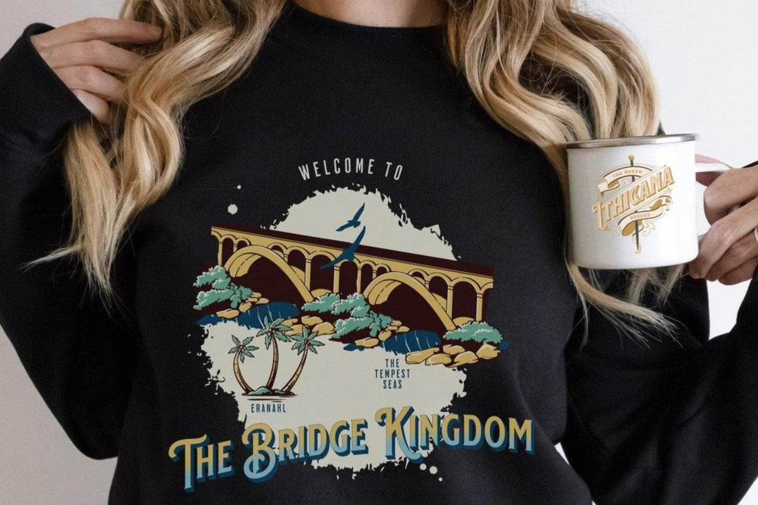 Woman holding Ithicana mug, wearing The Bridge Kingdom fantasy book merch sweatshirt with illustrated bridge design.