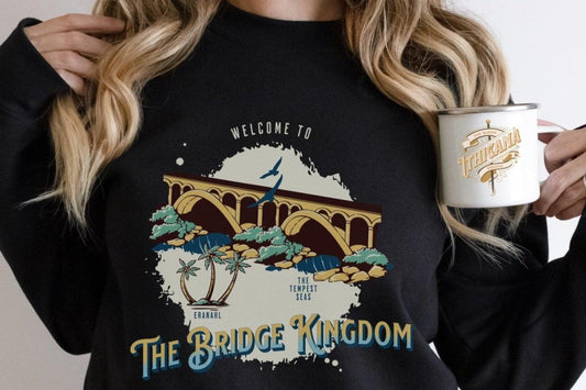 Woman holding Ithicana mug, wearing The Bridge Kingdom fantasy book merch sweatshirt with illustrated bridge design.