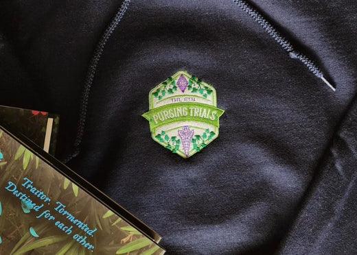Green Purging Trials embroidered patch on black fabric, next to an open fantasy book