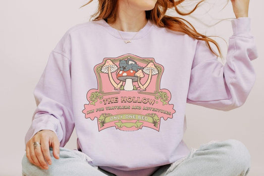 Woman wearing pastel sweatshirt with mushroom and cat graphic, reading 'The Hollow Inn for Travelers and Adventures, Only One Bed.'