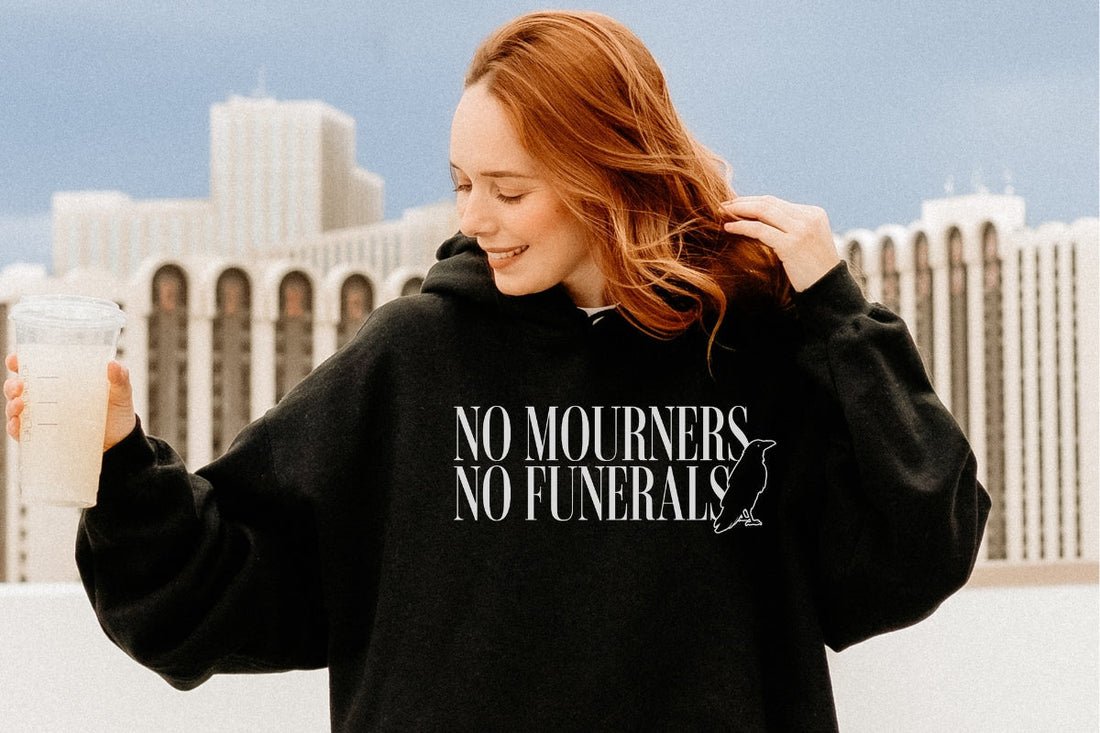 Woman wearing black "No Mourners No Funerals" fantasy book hoodie, holding a drink outdoors.