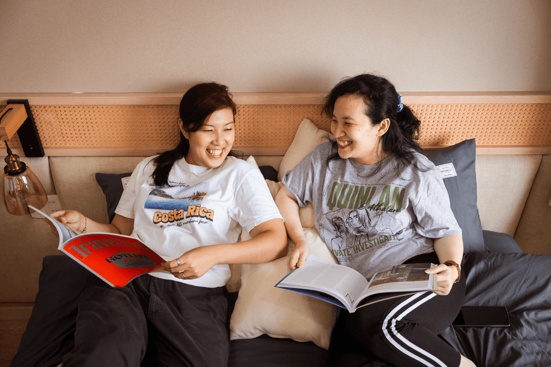 Two women smiling and reading books together on a cozy bed, hosting a relaxed book club.