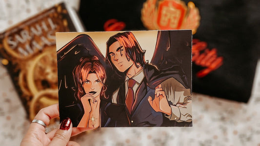 Hand holding a bookish art print of two fantasy characters with dark wings, surrounded by Crescent City novel and merch.