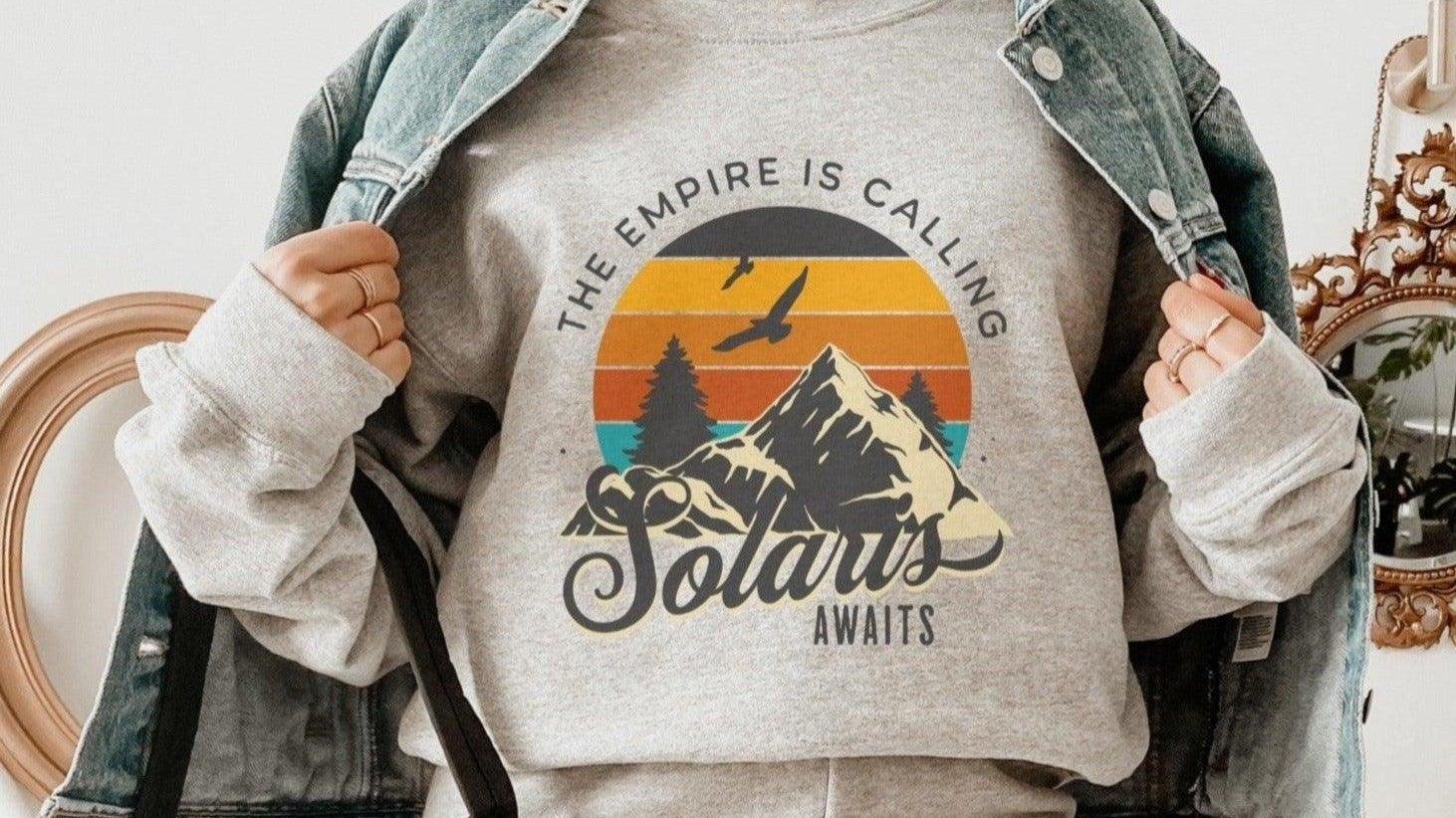 Person wearing a cozy bookish sweatshirt with mountain, trees, sunset, and text design