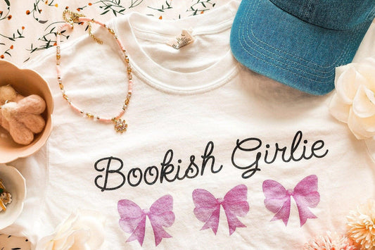 White t-shirt with 'Bookish Girlie' text and pink bows, styled with a blue denim cap, floral necklace, and pastel flowers.