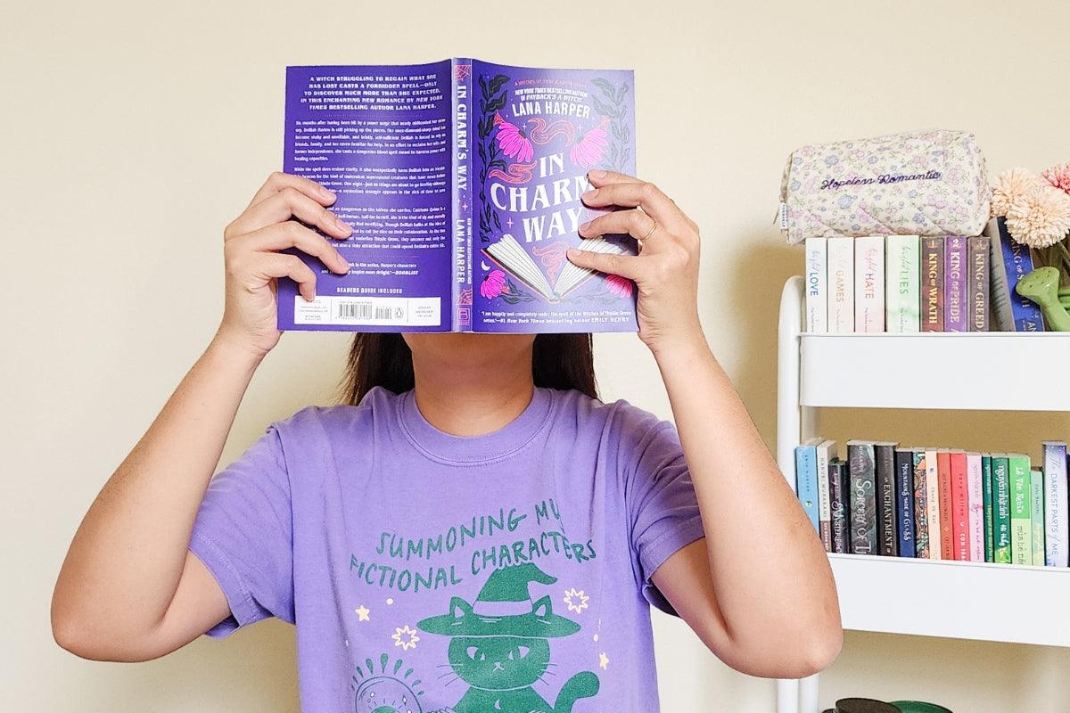 Person in purple shirt holding open book in front of face, bookshelf with novels in background