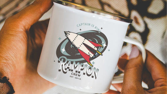White enamel mug with spaceship and 'Rampion Crew' illustration held in hands