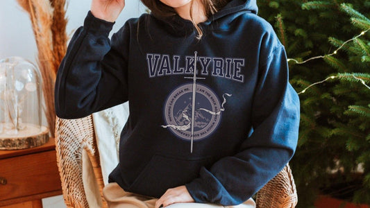 Person wearing a black Valkyrie hoodie with sword and mountain graphic, cozy indoor setting