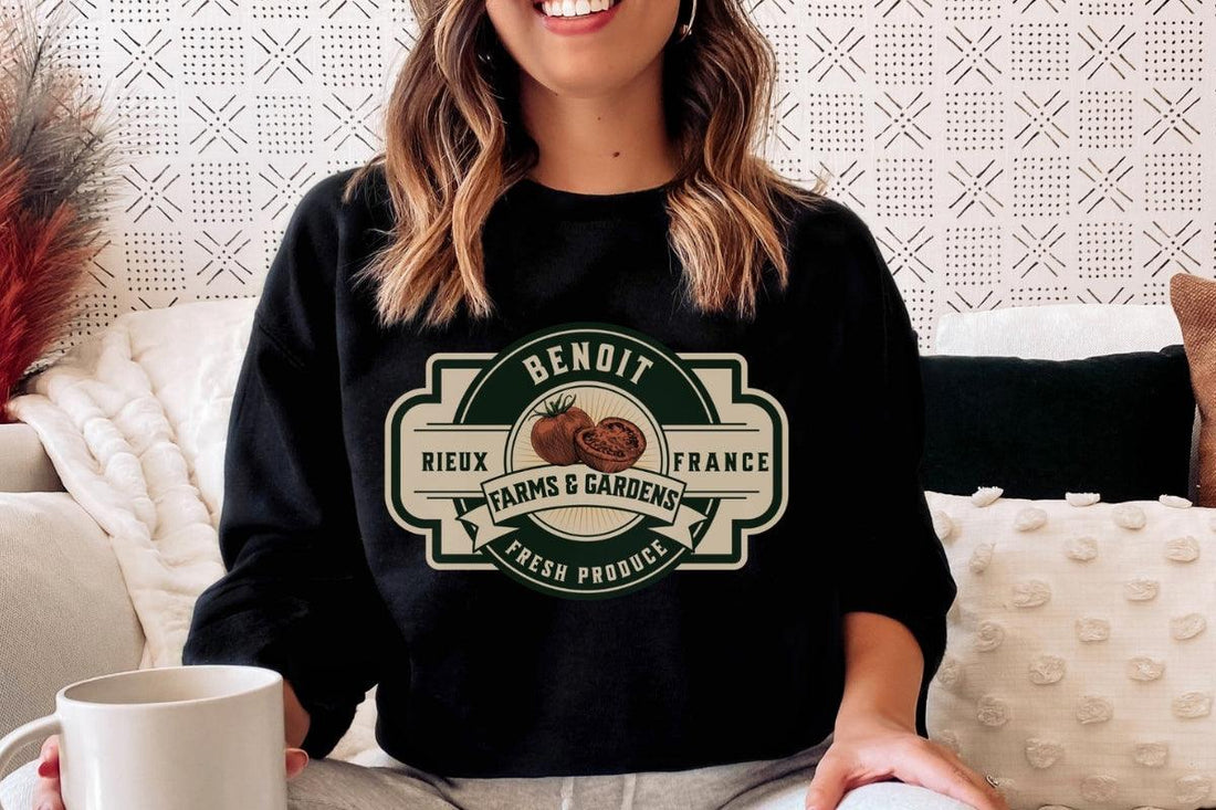 Woman sitting on cozy couch wearing black Benoit Farms & Gardens sweatshirt with coffee mug