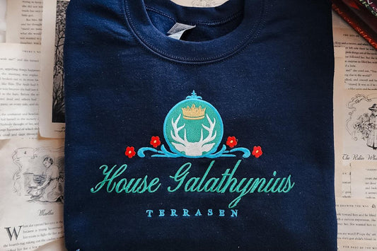Navy sweatshirt with House Galathynius Terrasen crest and floral embroidery on book pages.