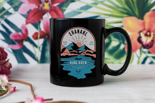 Black ceramic mug with a colorful mountain and lake graphic, surrounded by tropical flowers