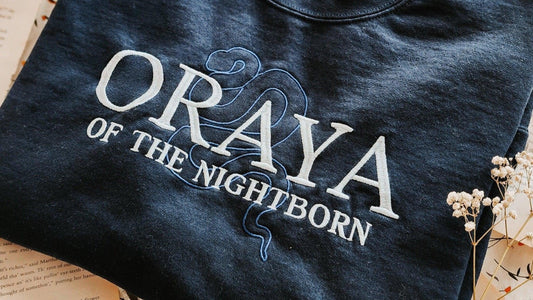 Navy crewneck sweatshirt with 'Oraya of the Nightborn' embroidered, a snake outline, and dried flowers.