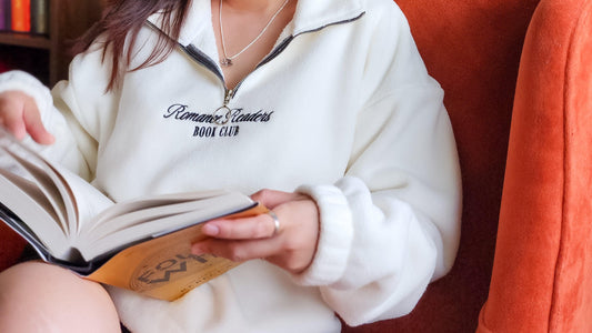 Person in white fleece Romance Readers Book Club pullover reading a book on an orange chair