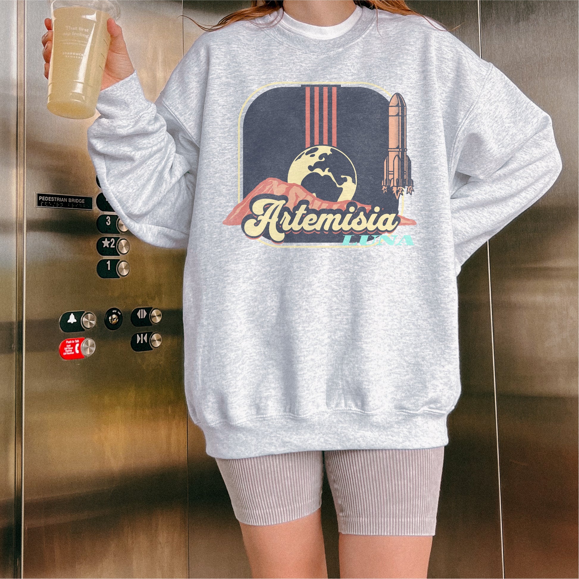 Woman wearing gray Artemisia Luna sweatshirt with rocket and moon graphic, holding drink in elevator