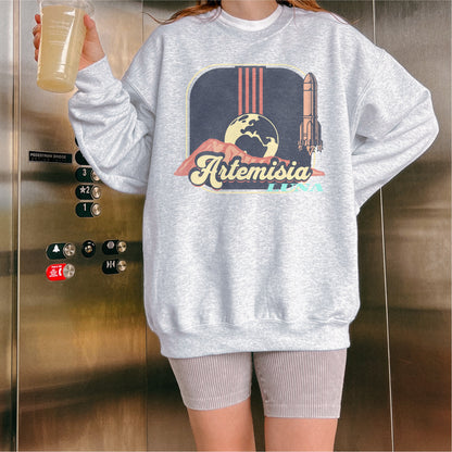Woman wearing gray Artemisia Luna sweatshirt with rocket and moon graphic, holding drink in elevator