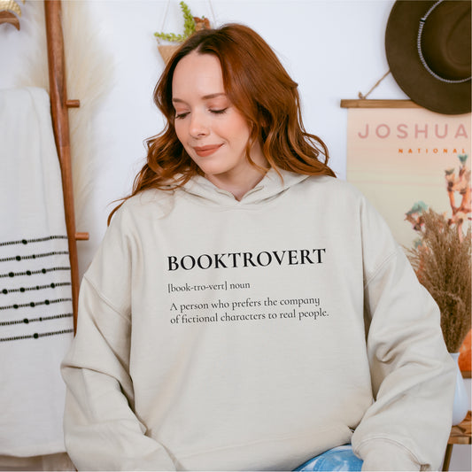 Woman wearing beige hoodie with 'BOOKTROVERT' book lover definition design, cozy indoor setting