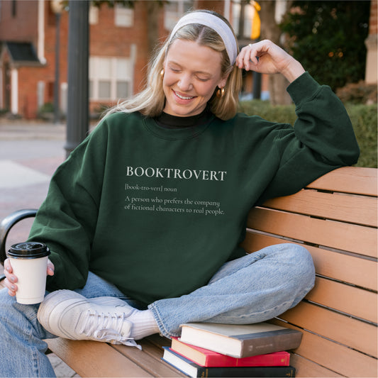 Smiling woman in green 'Booktrovert' sweatshirt sitting on a bench with books and coffee.