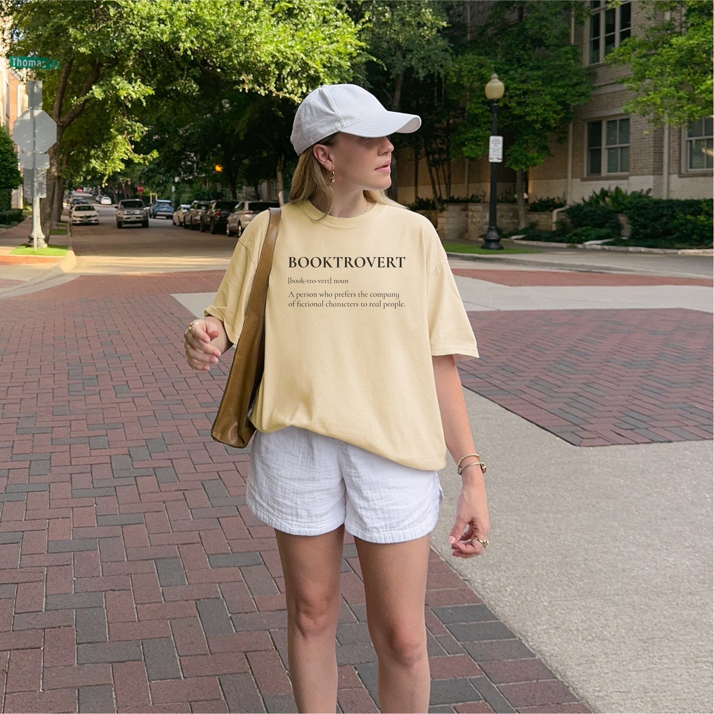 Woman wearing a beige BOOKTROVERT t-shirt and white shorts walking outdoors.