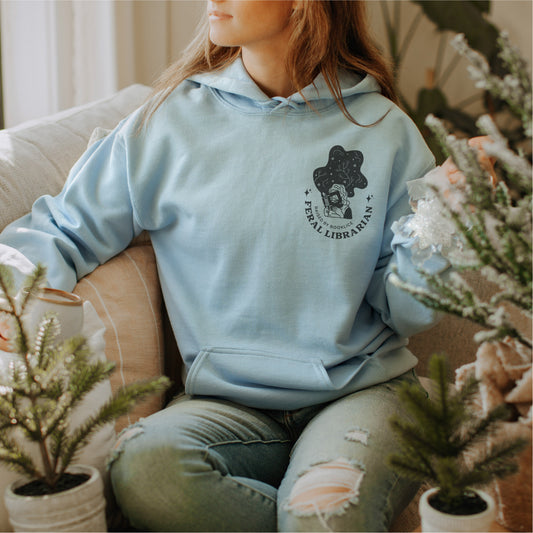 Woman wearing light blue Feral Librarian hoodie with graphic, sitting on cozy sofa