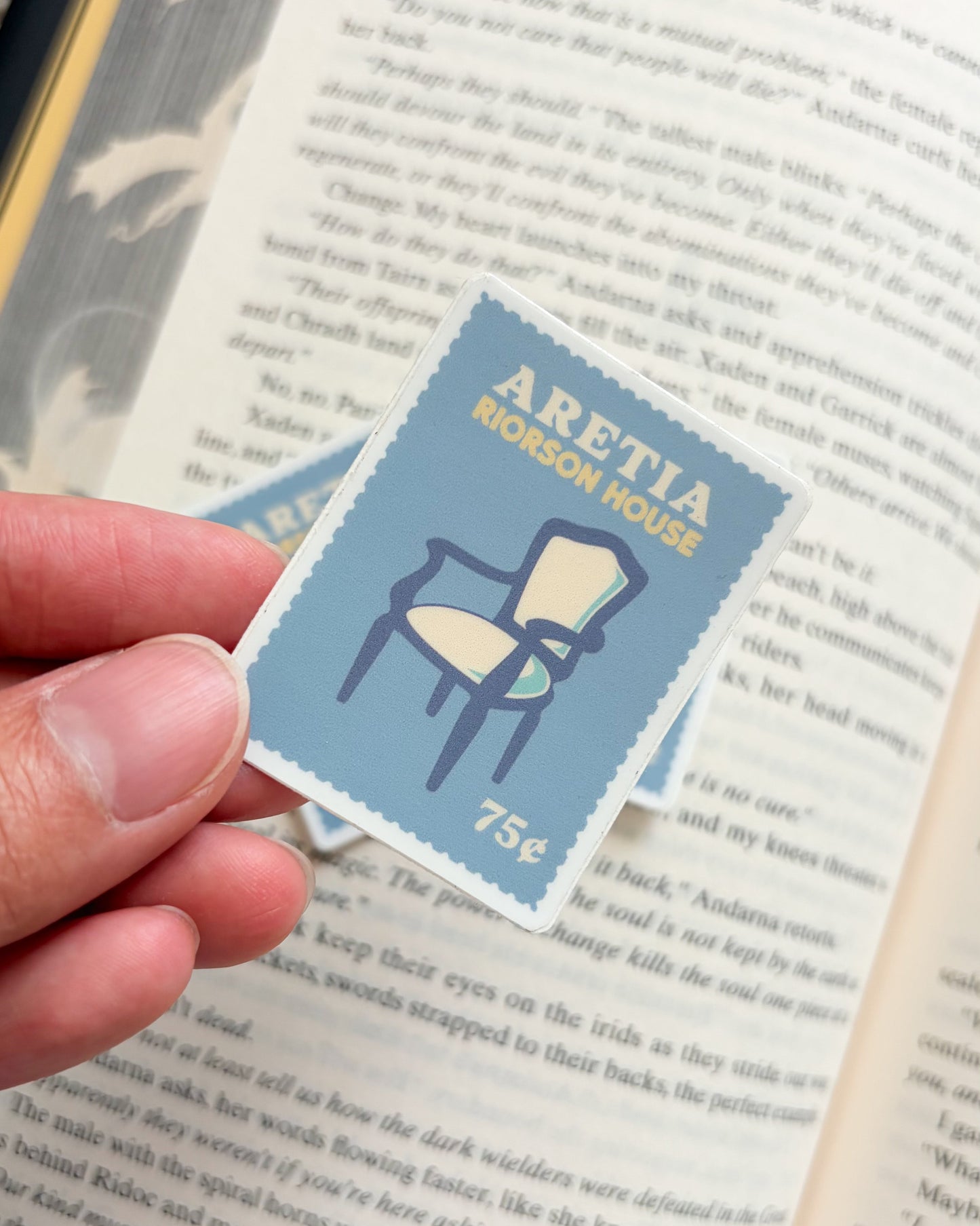 Small blue and white stamp with 'Aretia' and 'Riorson House' text held over an open book.