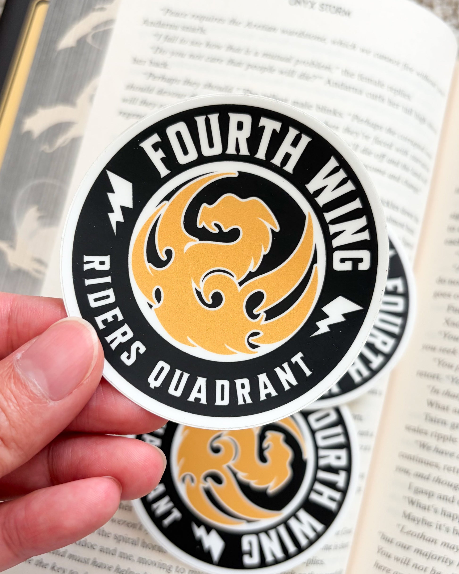 Sticker with 'Fourth Wing Riders Quadrant' design held over an open book.