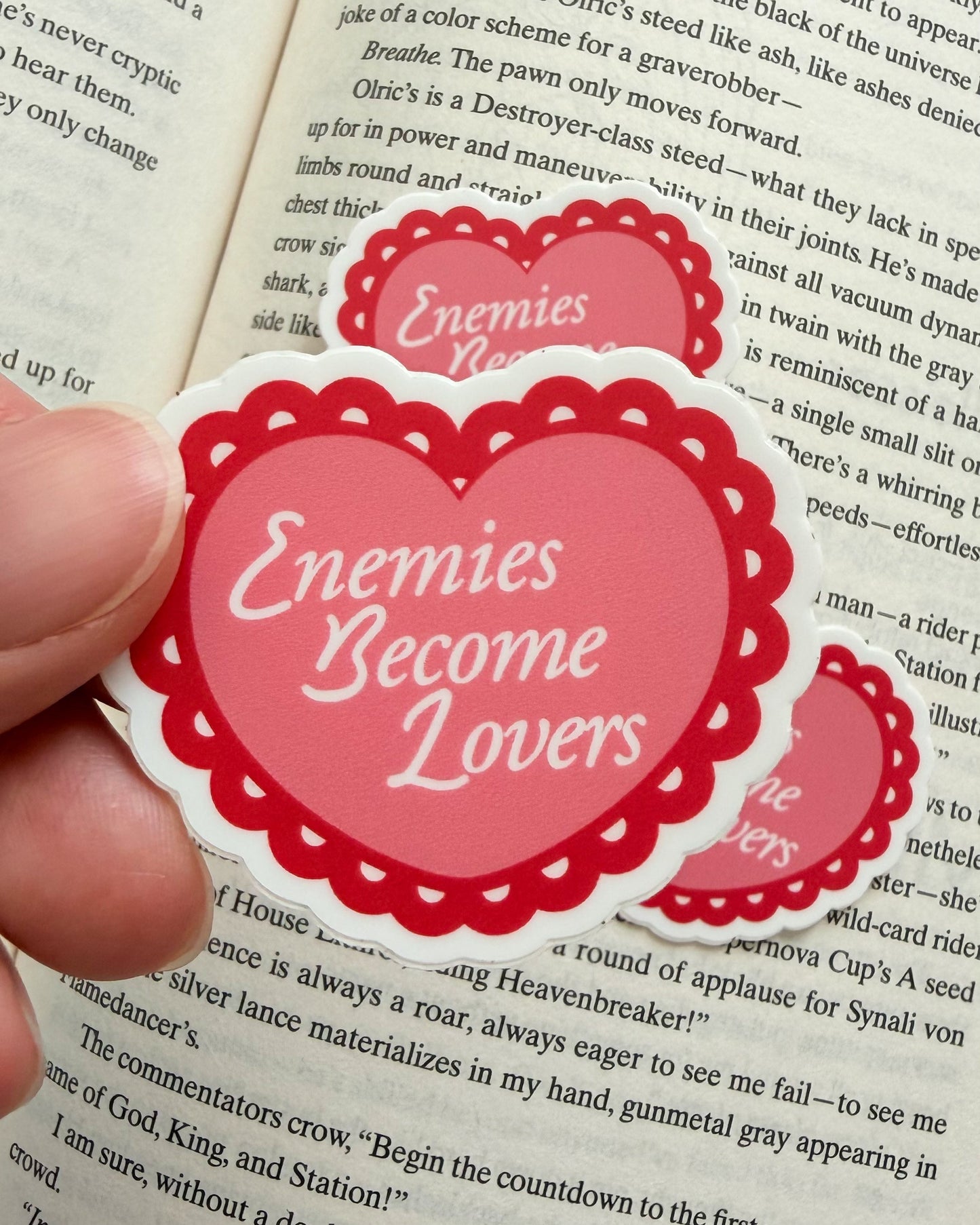 Heart-shaped sticker with 'Enemies Become Lovers' text held over an open book.