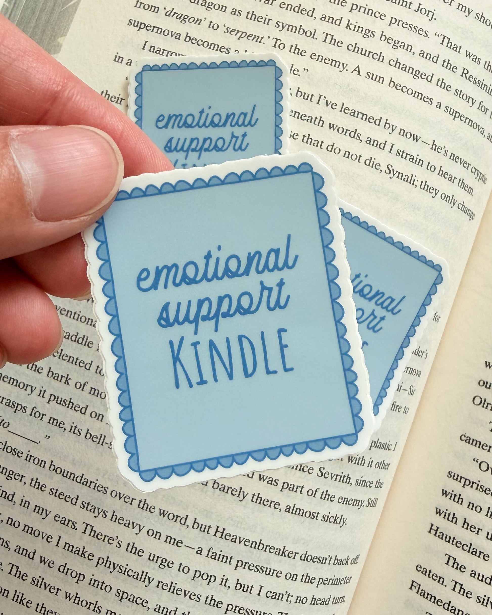Hand holding a blue 'emotional support Kindle' sticker over an open book