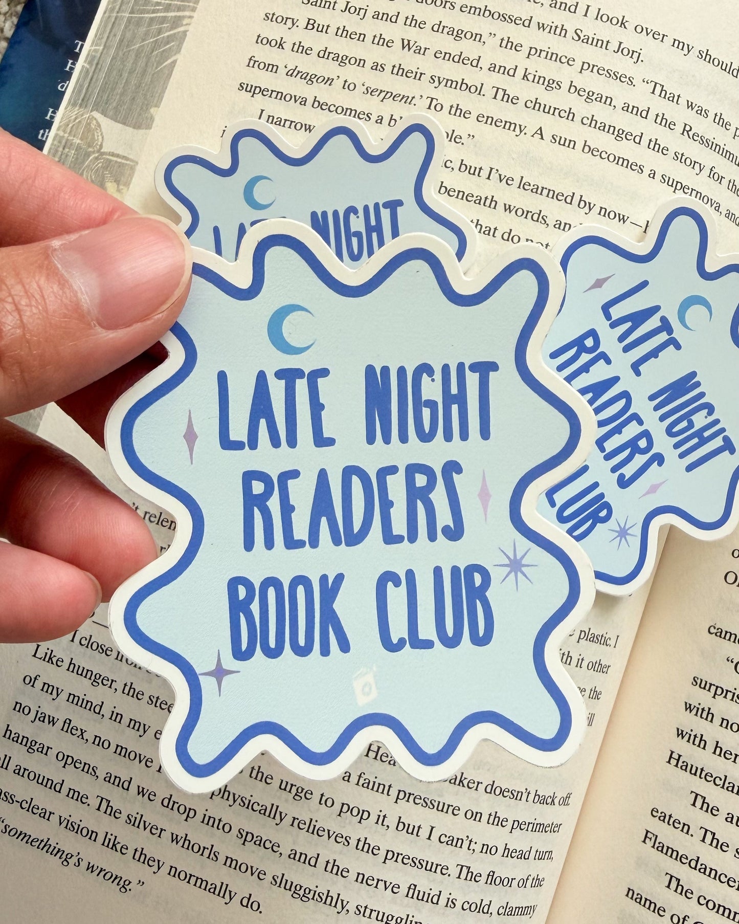 Hand holding a 'Late Night Readers Book Club' sticker over an open book