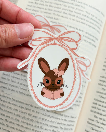 Hand holding a decorative bunny sticker with a bow over an open book