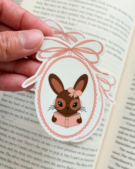 Hand holding a decorative bunny sticker with a bow over an open book
