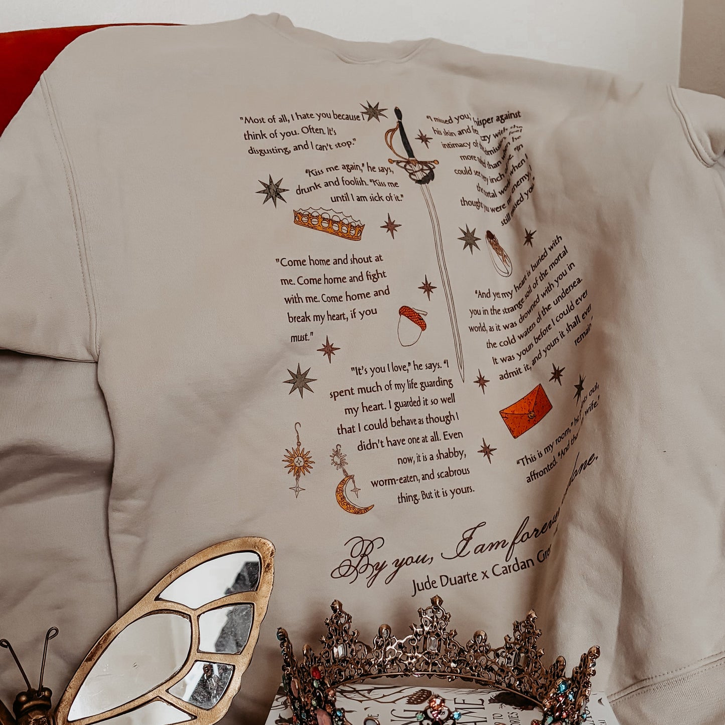 Beige bookish sweatshirt with literary quotes and fantasy graphics, displayed with crowns and mirror.