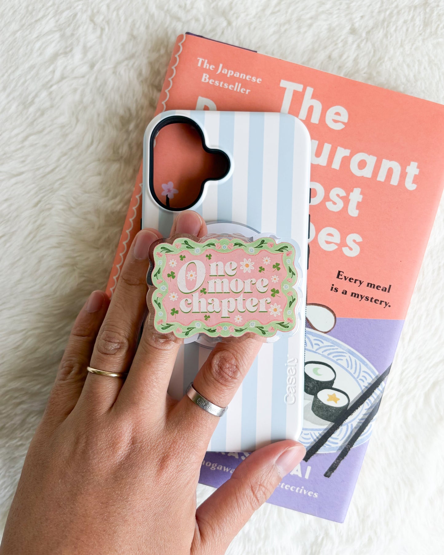 A hand holds a light blue and white striped phone case with a pink and green 'One More Chapter' cookie-shaped pop socket, positioned over a book on a white faux fur background.