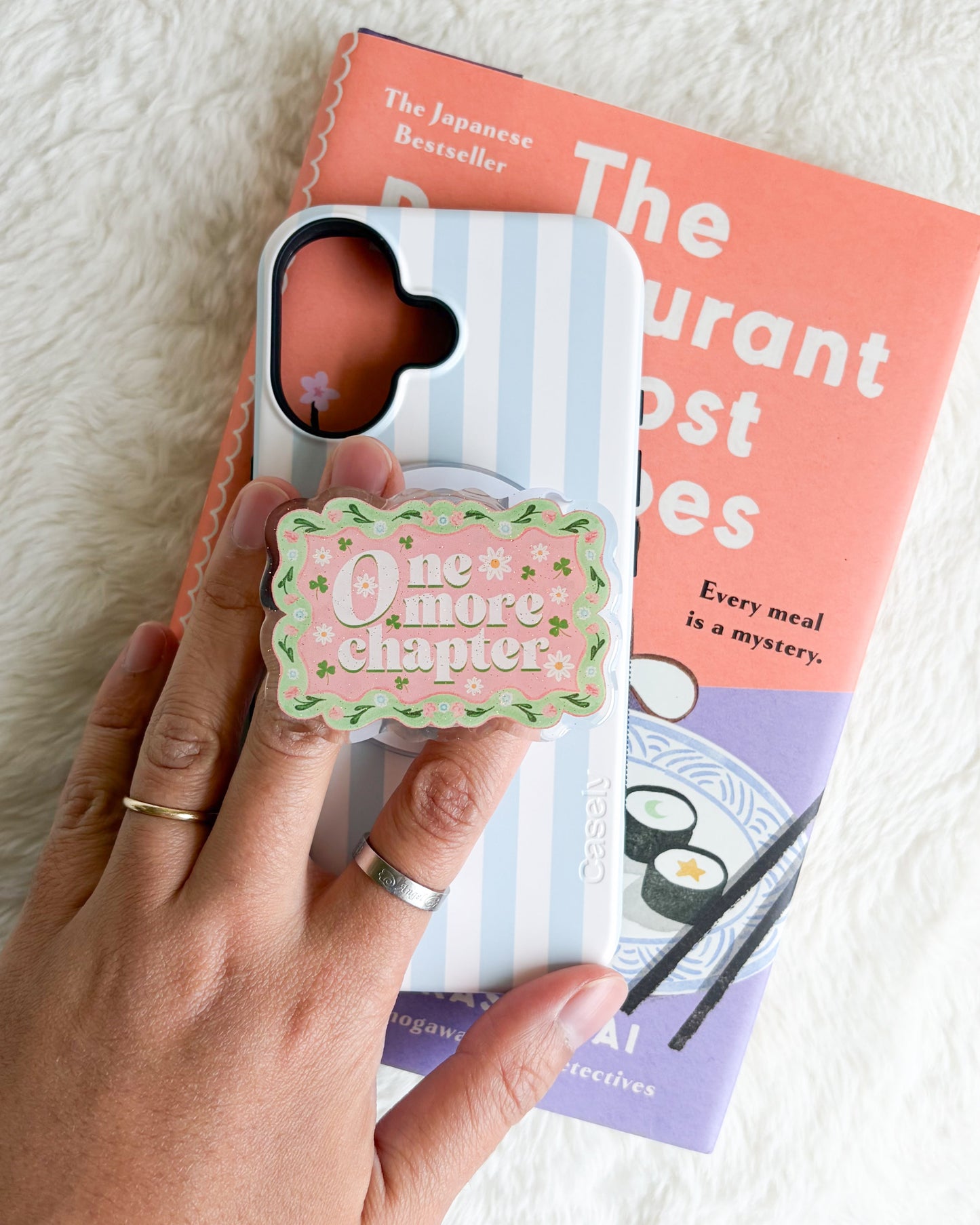 A hand holds a light blue and white striped phone case with a pink heart-shaped camera cutout and a decorative pop socket that reads 'One more chapter'. The case is styled over a pink and purple book on a white faux fur background.
