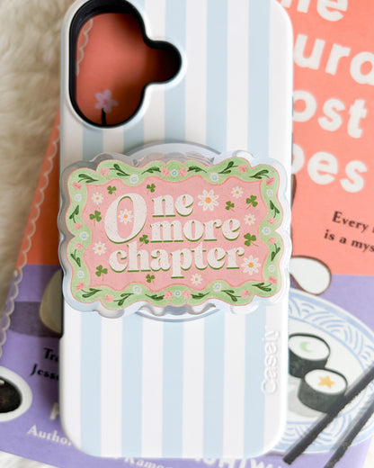 A phone case with a light blue and white vertical stripe pattern, featuring a central pink decorative grip with the text 'One more chapter' in white script, surrounded by small green leaves. The case is displayed on top of books on a white textured surface.