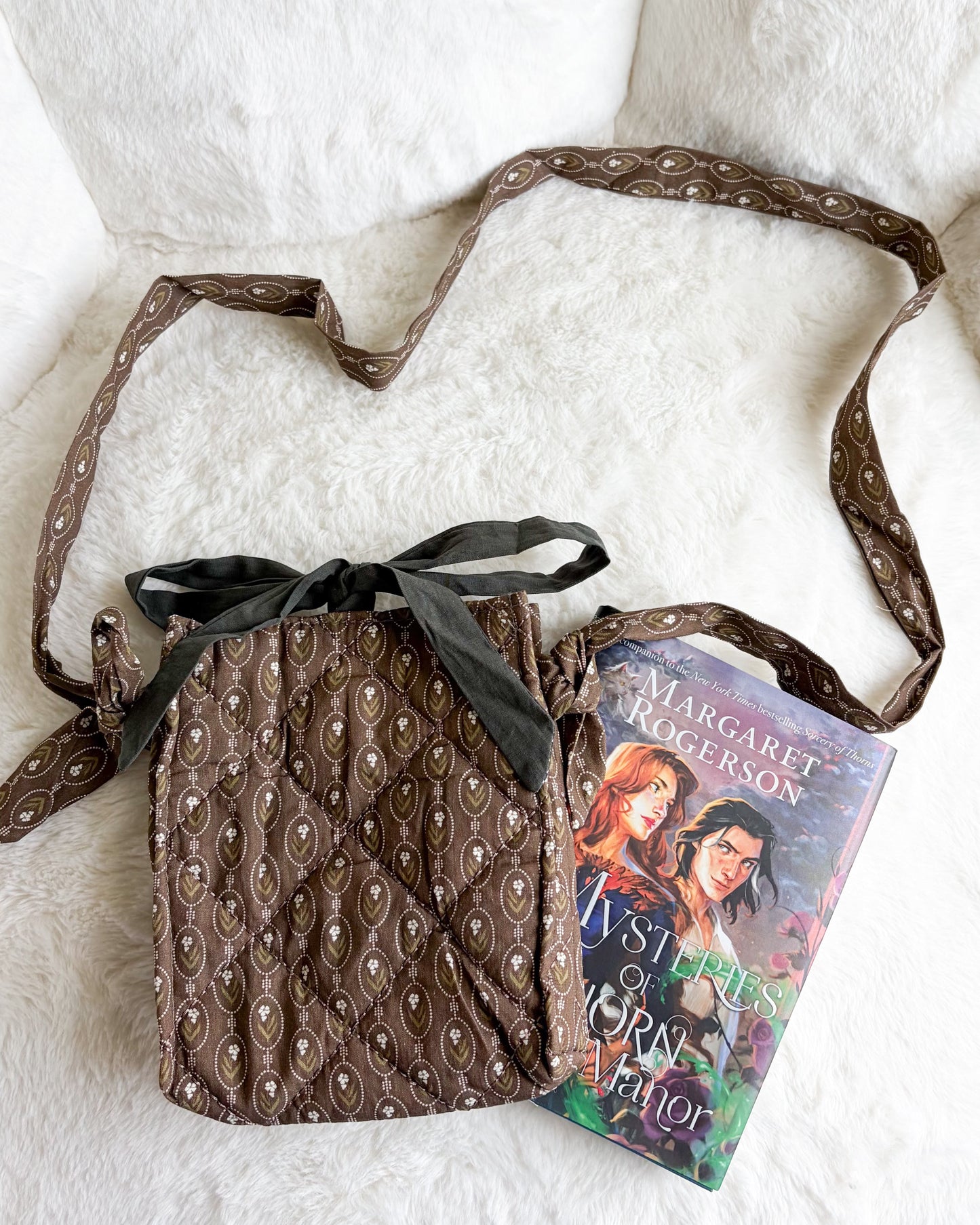 A brown patterned fabric crossbody bag with a black ribbon bow, styled on a white faux fur surface next to a book.