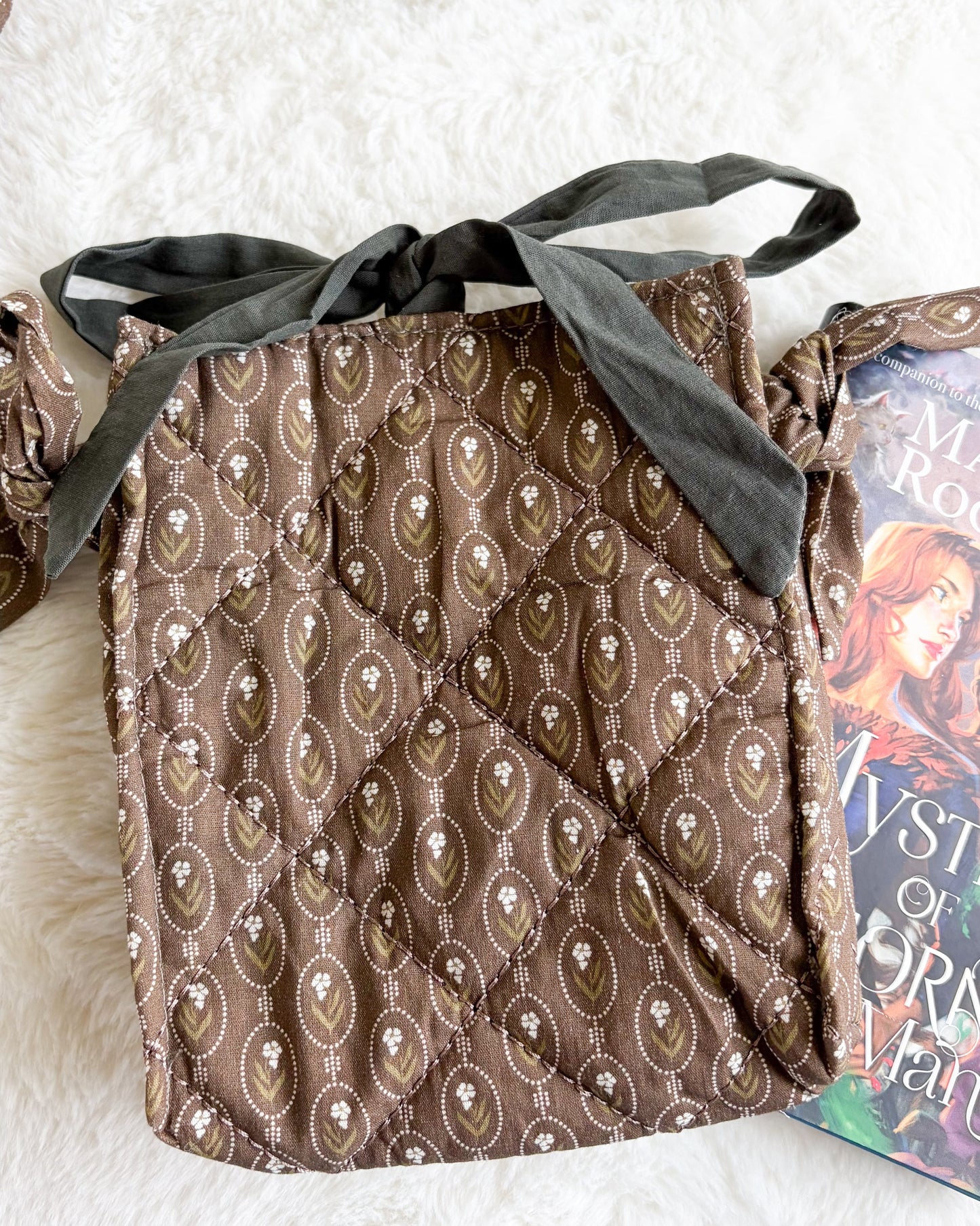 A brown quilted fabric bag with an allover paisley pattern, featuring a dark gray fabric ribbon tied in a bow at the top, laid flat on a white faux fur surface next to a book.
